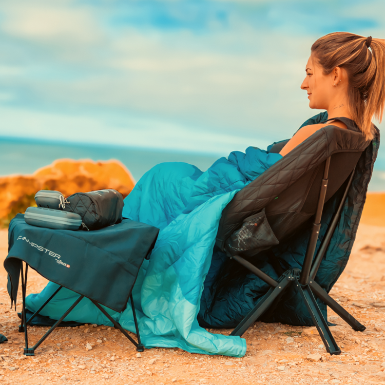 A woman sitting on a coastal cliffside in the Campster 2 camping chair with a camping blanket and a foldable camping table with a microfiber towel and travel accessories on the top.
