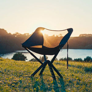 A travel sized foldable chair, the Campster 2, sitting in the grass in front of a lake with a sunset peering through the lightweight mesh backing. 